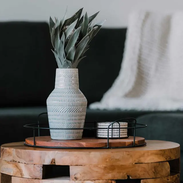 Vernon Court Vase with dried leaves on round wooden table, black metal tray, two coasters; dark sofa and white blanket in background.