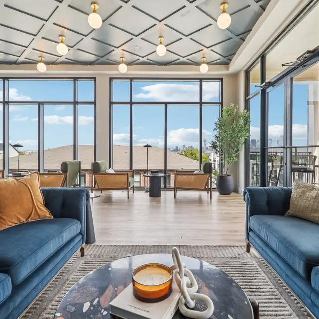 A spacious modern living room with large windows showcasing a view. There are two blue sofas adorned with cushions, a coffee table at the center, and stylish lighting fixtures on the ceiling.