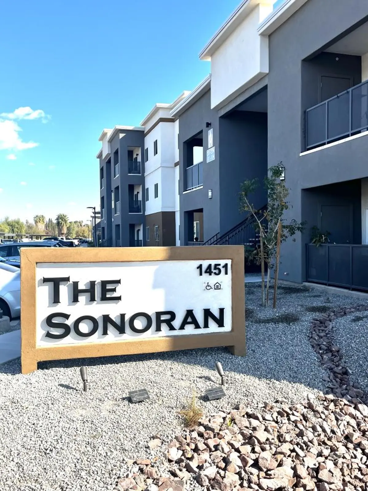 Monument sign marked The Sonoran and 1451 beside gravel and buildings.