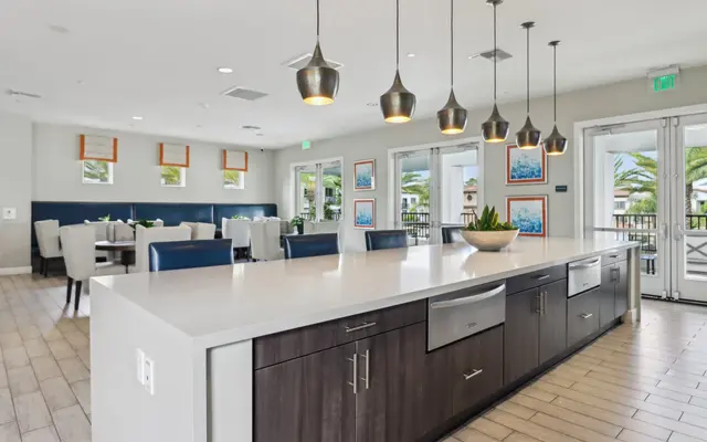 A modern kitchen with a large countertop island, elegant pendant lighting, and a seating area. Large windows allow natural light to illuminate the space, decorated with plants and artwork.