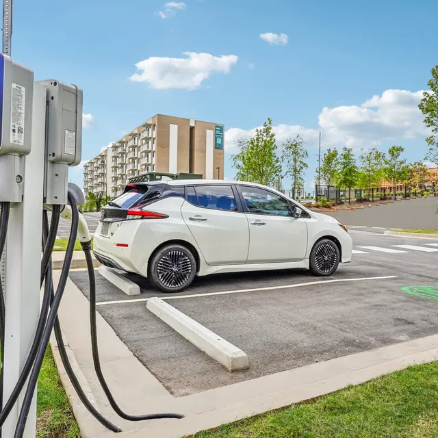 An electric vehicle charging station with a white car parked next to it. In the background, apartment buildings and trees are visible under a blue sky with clouds.