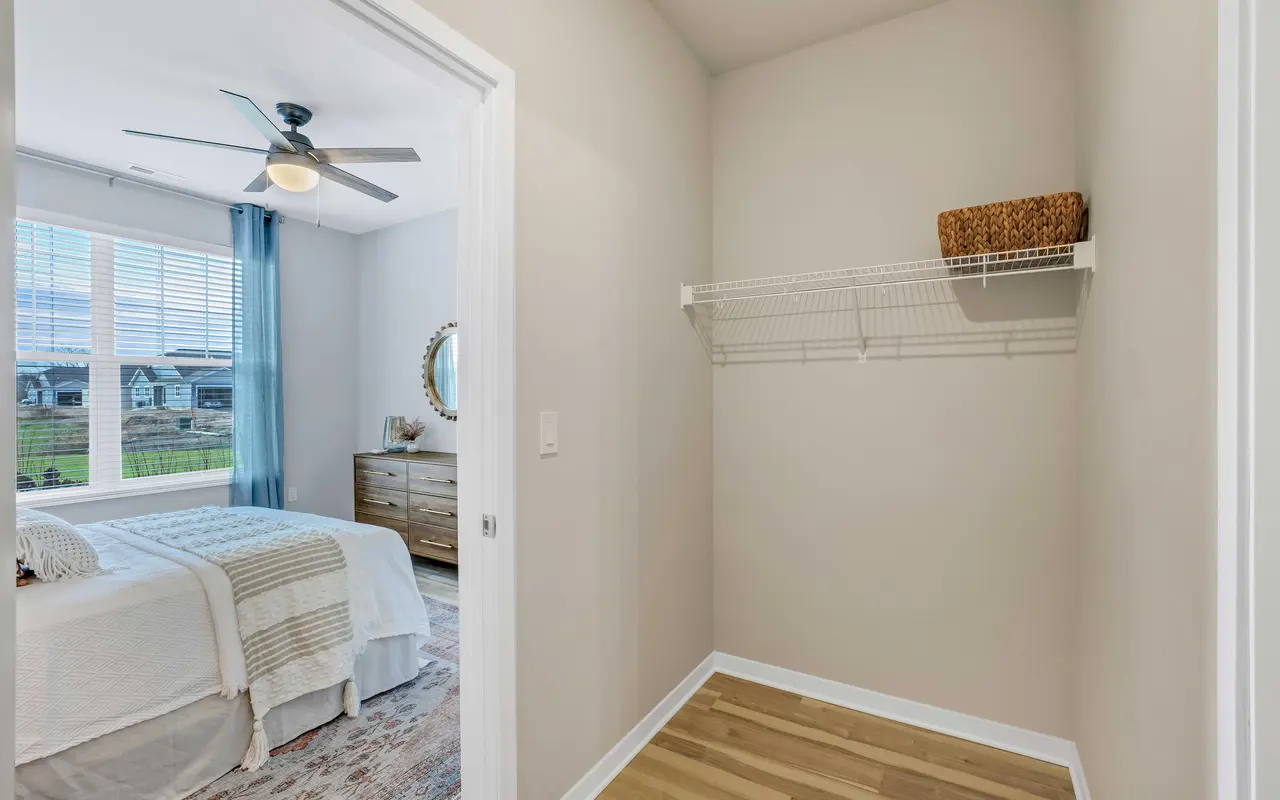 Inside view of a modern bedroom with a doorway leading to a closet space. The room features a bed with a light-colored bedspread, a dresser, and large windows with blue curtains. A mirror hangs on the wall, and the floor is made of light wood.