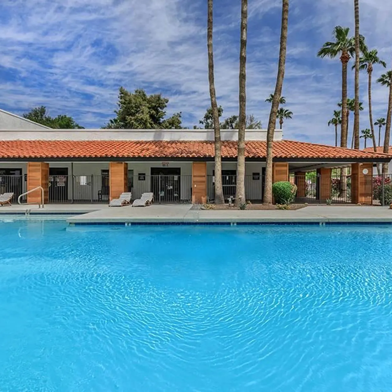 A bright swimming pool in front of a building with a tiled roof, surrounded by palm trees and greenery under a blue sky.