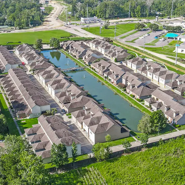 Aerial view of a residential housing complex with a central waterway, surrounded by greenery and a road in the background.