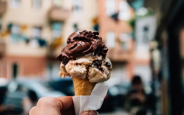 A hand holding an ice cream cone with two scoops: one chocolate and one vanilla with chocolate chunks, set against a blurred background of a street scene.