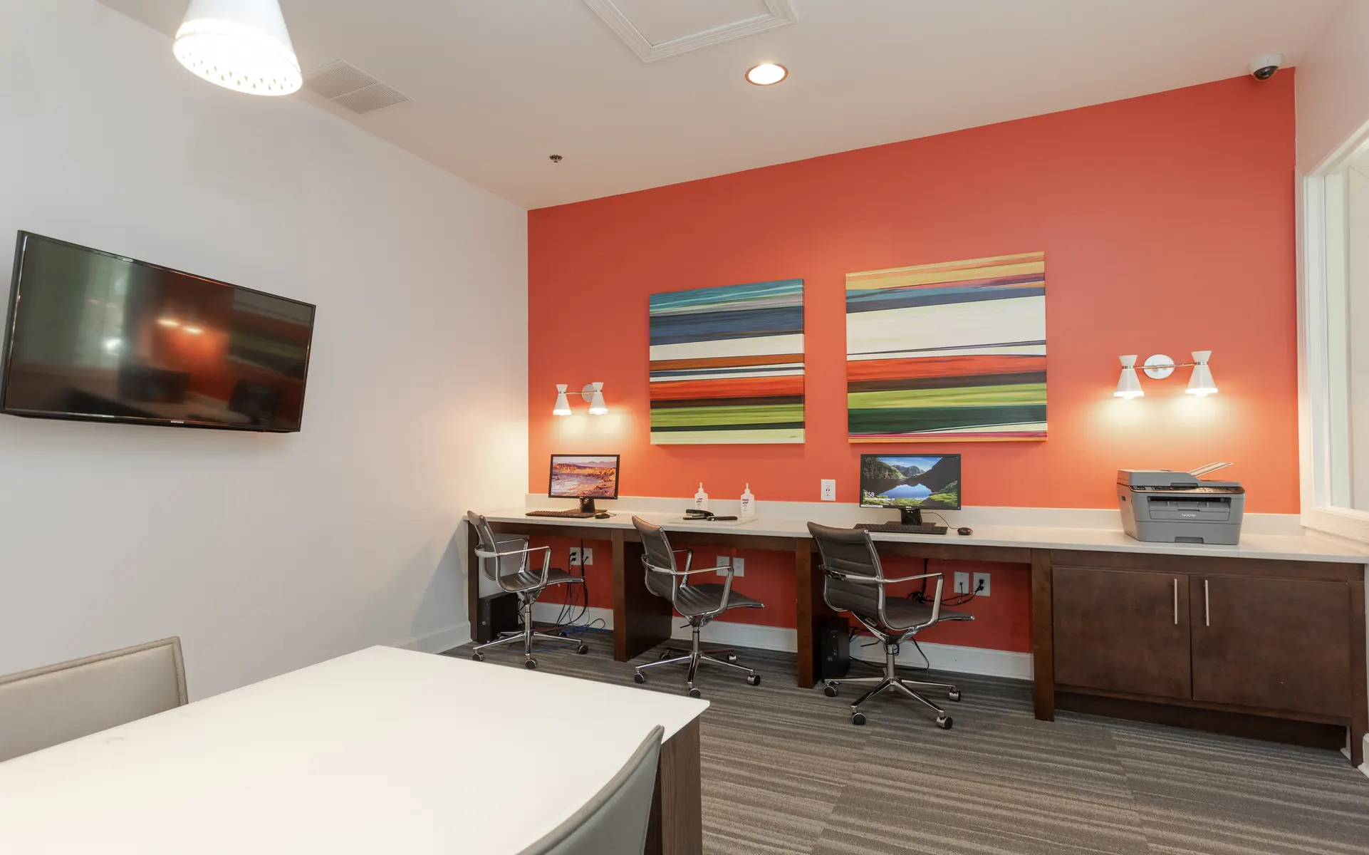 Modern office interior with a bright orange accent wall, two workstations with computers, a printer, decorative wall art, and a television mounted on the wall.