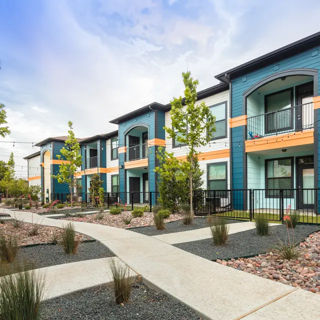 A modern apartment complex with blue and white exterior, featuring multiple balconies, surrounded by landscaped gardens with pathways and small plants.