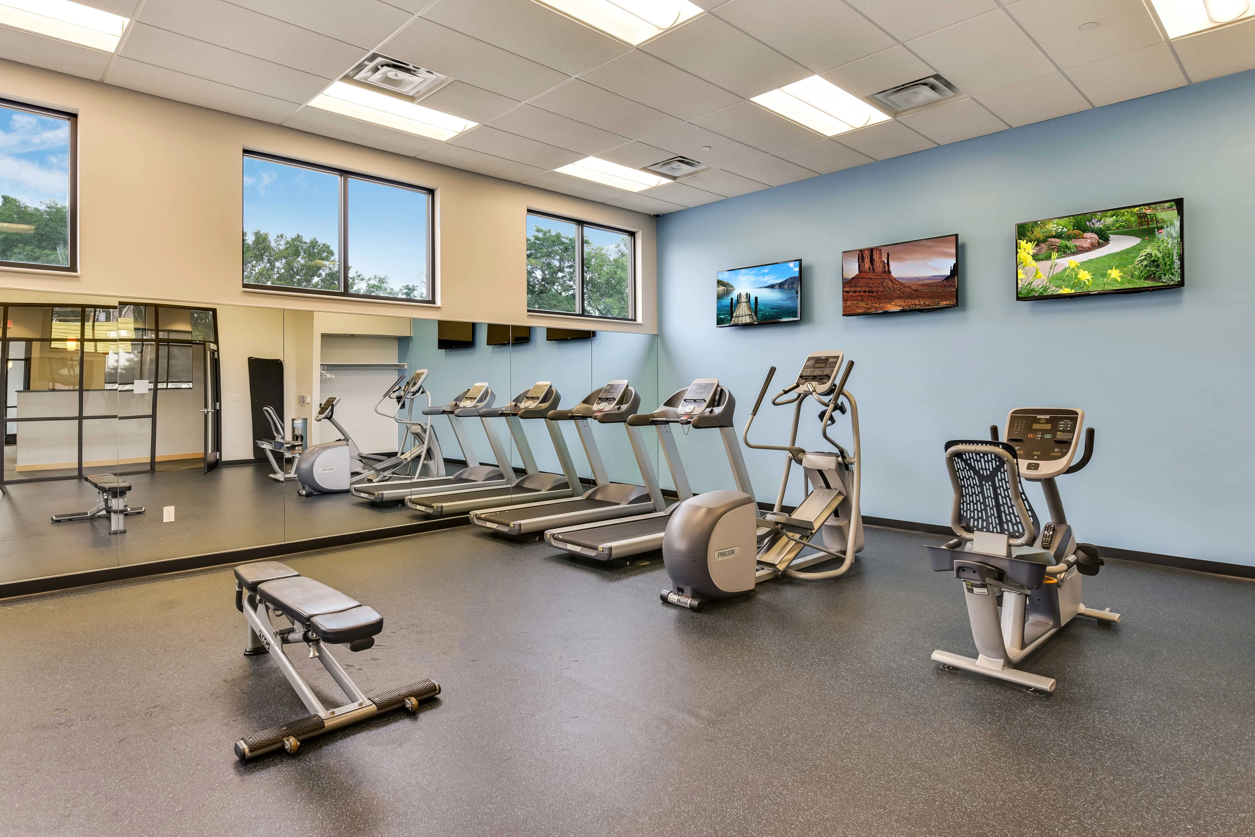 Modern Gym Interior A modern gym interior featuring multiple treadmills, an elliptical machine, and a stationary bike. Large windows bring in natural light, and there are flat-screen TVs mounted on the wall displaying scenic landscapes. The flooring is black rubber, and there's a bench in the foreground.