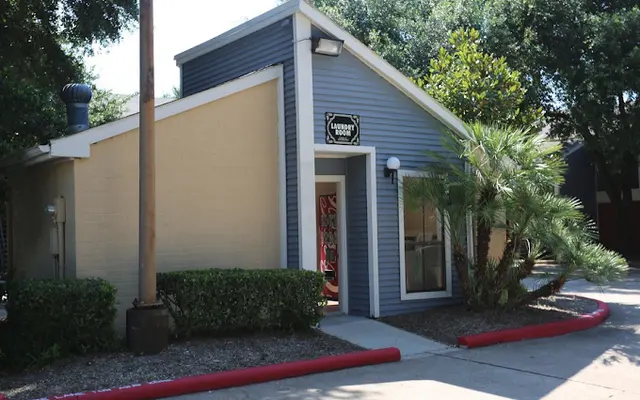 A small, modern building with a pitched roof, painted in gray and cream, featuring a sign that reads 'Lassiter Lodge.' The entrance is framed by large windows and surrounded by shrubs and palm trees. A red curb lines the driveway, and there are green trees in the background.