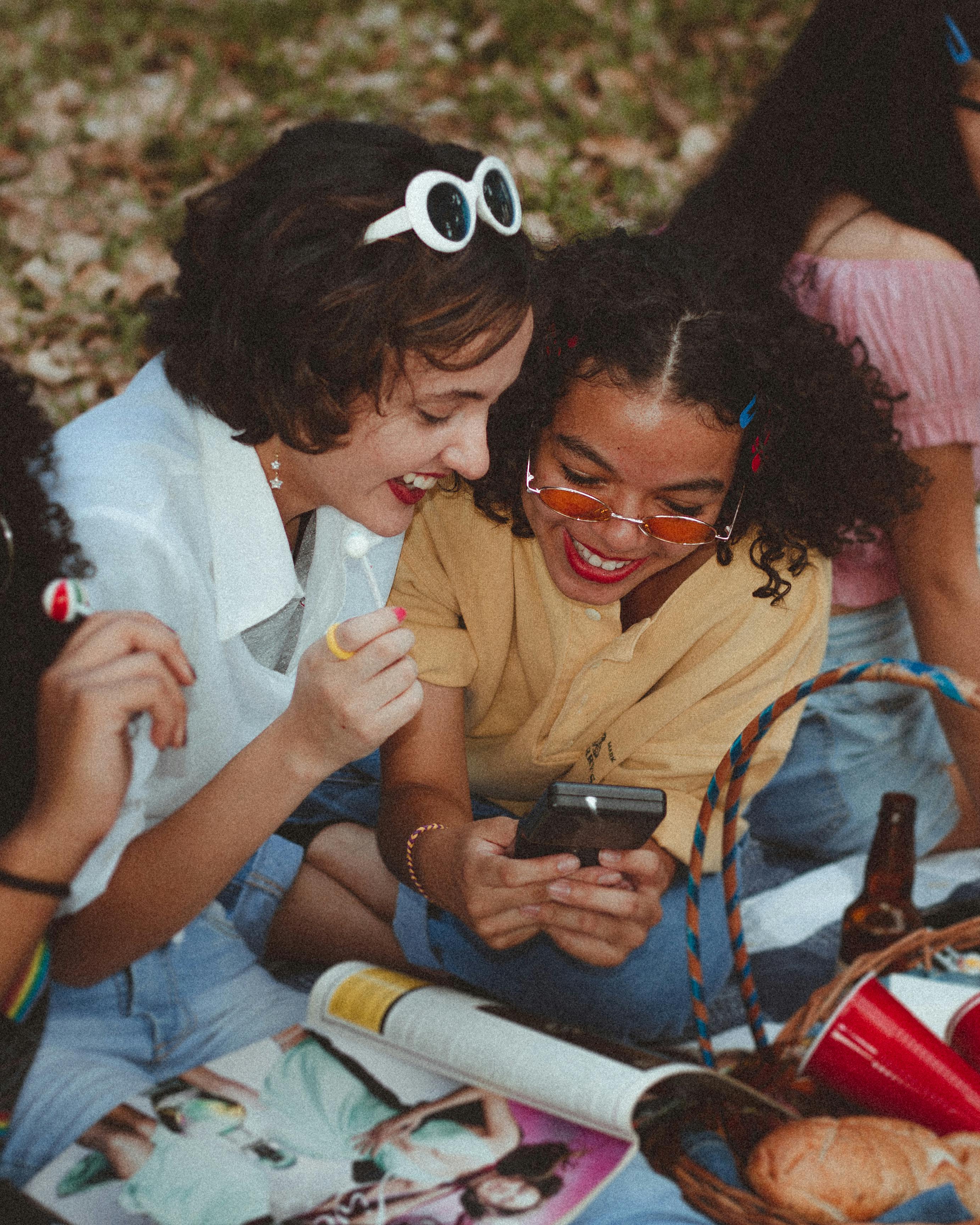 Friends Laughing Together Outdoors Two young women sitting on the ground enjoying a playful moment while looking at a phone. They are surrounded by snacks and magazines, laughing together in a casual outdoor setting.