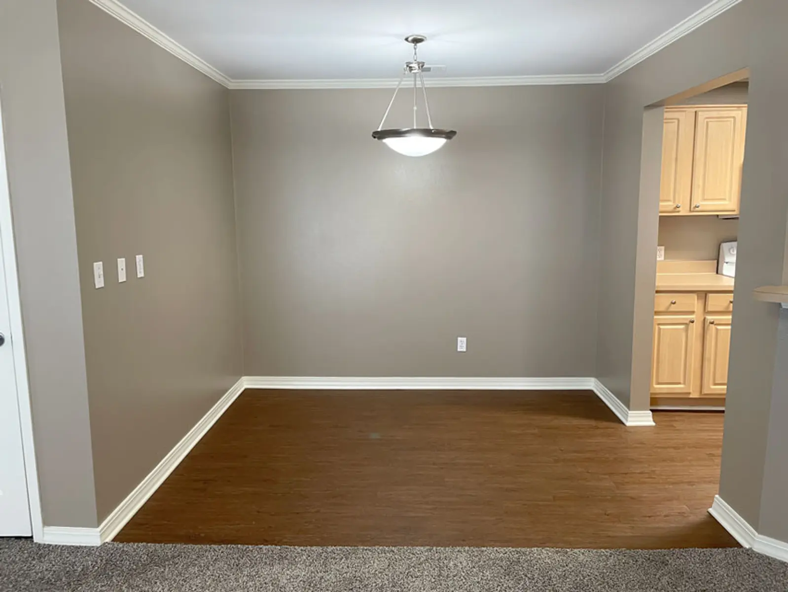 An empty dining area with a light fixture and a doorway leading to a kitchen.