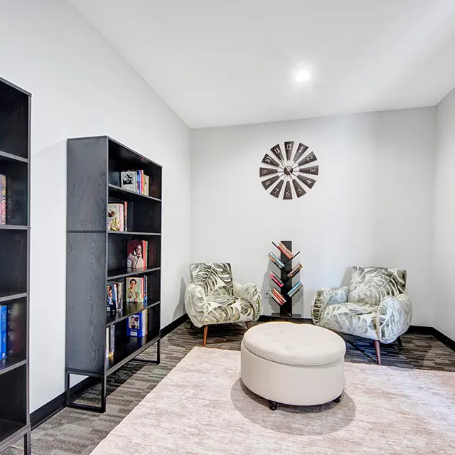 Cozy Reading Area A cozy reading space featuring two patterned chairs, a round ottoman, a bookshelf filled with books, and two decorative wall art pieces.