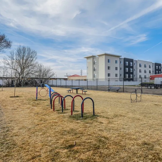 A grassy playground area with a small play structure and several bike racks, surrounded by a fence, with apartment buildings in the background.