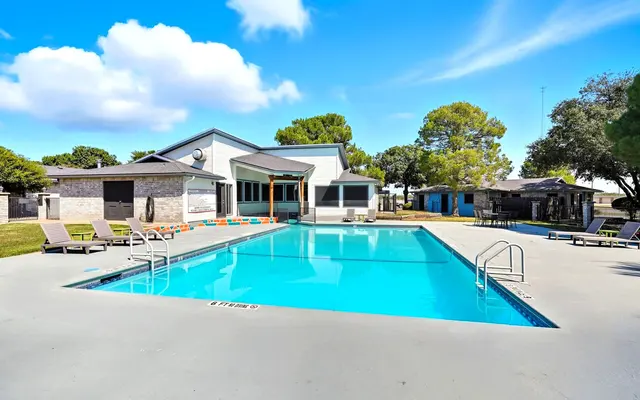 A bright outdoor swimming pool surrounded by lounge chairs and trees on a clear day.
