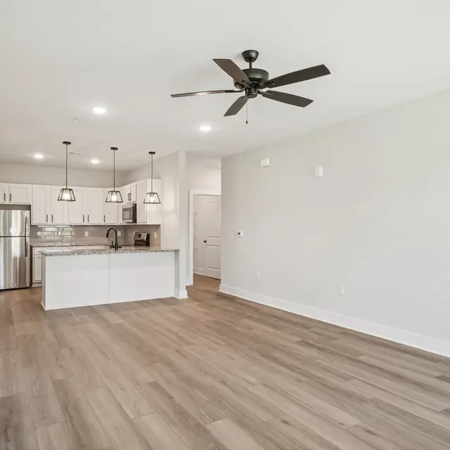 A modern open-concept living area featuring a kitchen with stainless steel appliances, a central island, and pendant lighting. The space has light-colored walls and wooden flooring, complemented by a ceiling fan. Natural light fills the room, giving it a bright and airy feel.