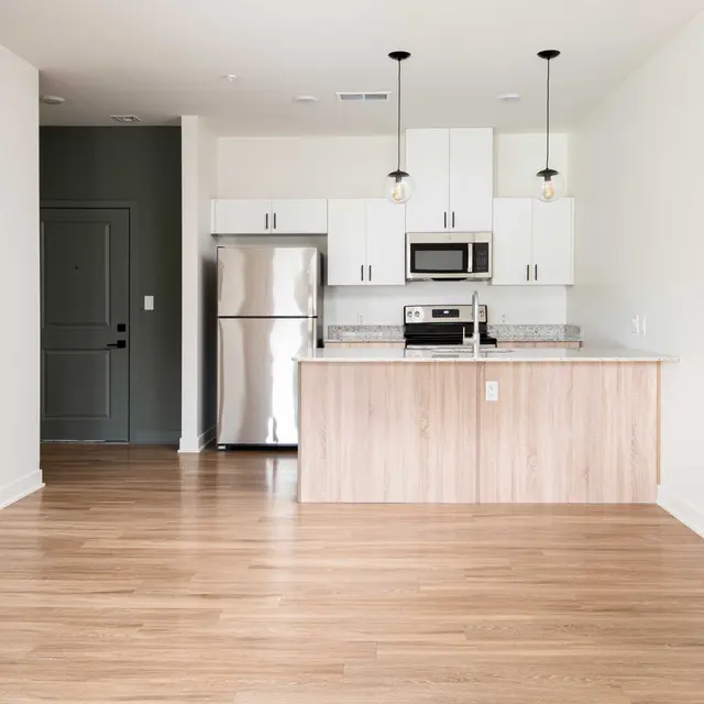 A modern kitchen with stainless steel appliances, white cabinets, and wooden flooring. The space features a bar-style countertop and pendant lighting, leading to a darkly painted door.