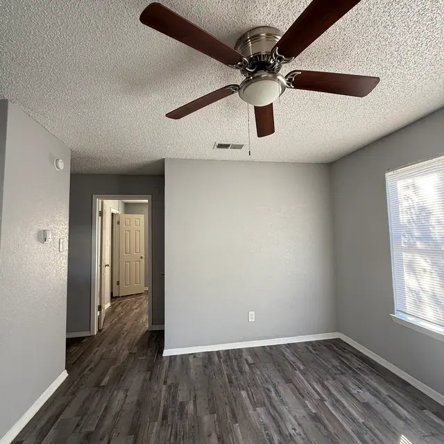 A spacious room featuring a ceiling fan, light gray walls, and laminate flooring. There is natural light coming through a window, and a doorway leading to another room can be seen in the background.