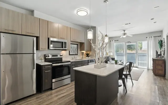 Modern kitchen with stainless steel appliances, an island, and a dining area, featuring natural light from large windows.