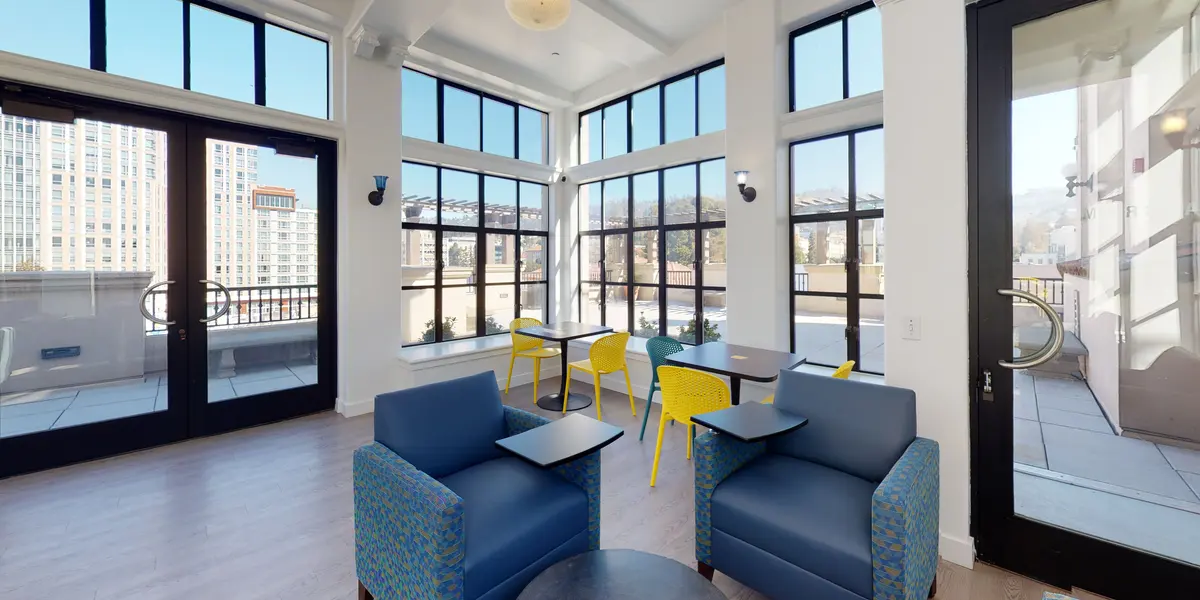 A bright lounge area with large windows offering views of a city. There are two blue chairs with a small table between them and several colorful yellow chairs and tables in the background.