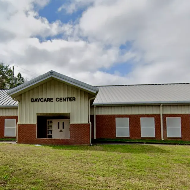 A single-story daycare center building with a large sign that reads 'DAYCARE CENTER'. The building features a light-colored façade with a gray metal roof. The surrounding area includes a grassy lawn and a few bare trees under a cloudy sky.
