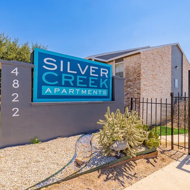 Sign for Silver Creek Apartments, displaying the name and address on a stone wall with a cactus in front, surrounded by a green lawn and trees under a clear blue sky.