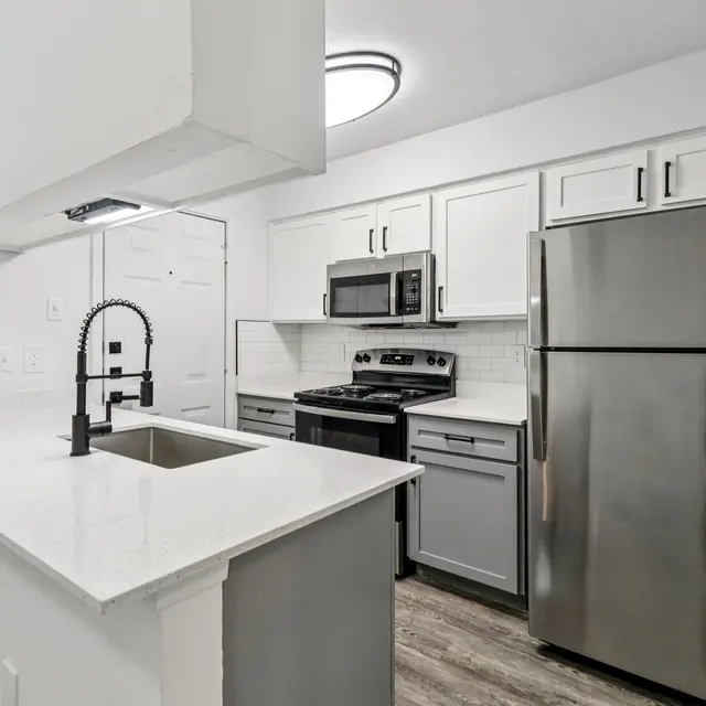 A modern kitchen featuring grey and white cabinetry, a stainless steel refrigerator, a gas stove, and a large island with a sink. The kitchen has a clean, sleek design with wood-look flooring and bright overhead lighting.