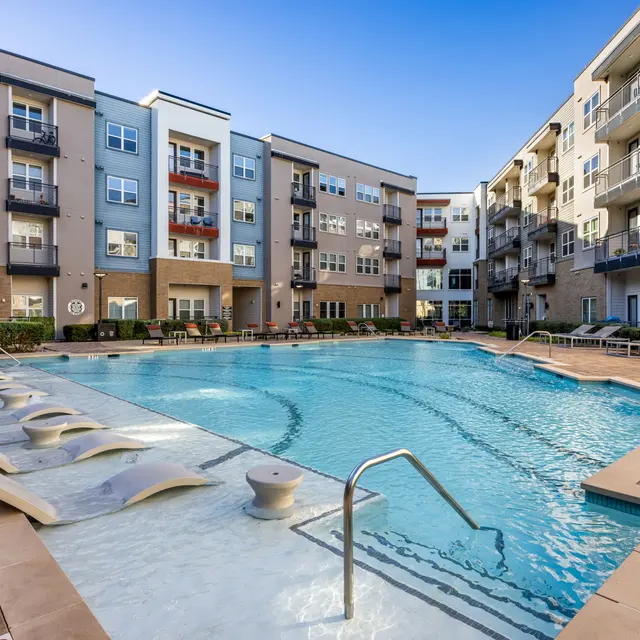 A modern apartment complex featuring a large swimming pool surrounded by lounge chairs and vibrant landscaping.