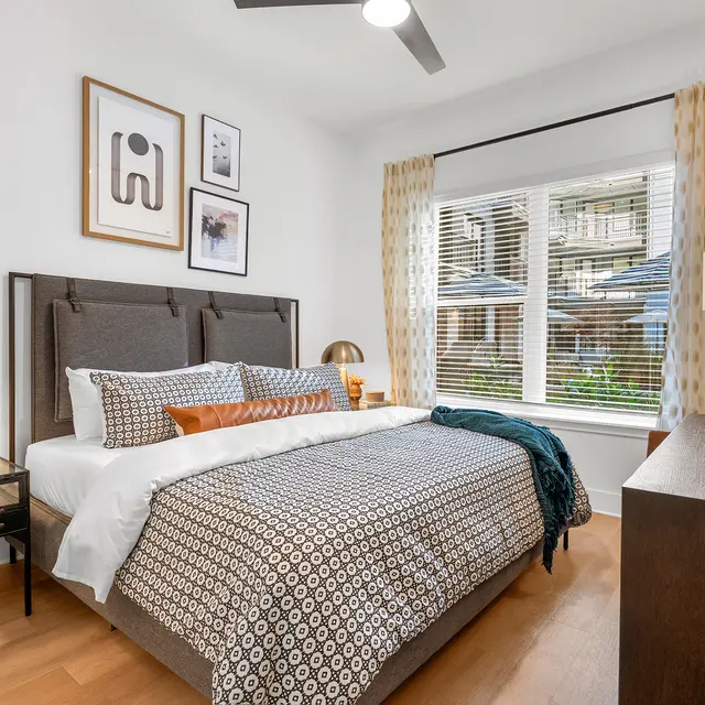 A modern bedroom featuring a queen-sized bed with patterned bedding, a bedside lamp, and a small nightstand. Bright window light enters through sheer curtains, illuminating the room with decorative wall art above the headboard and a wooden dresser nearby.