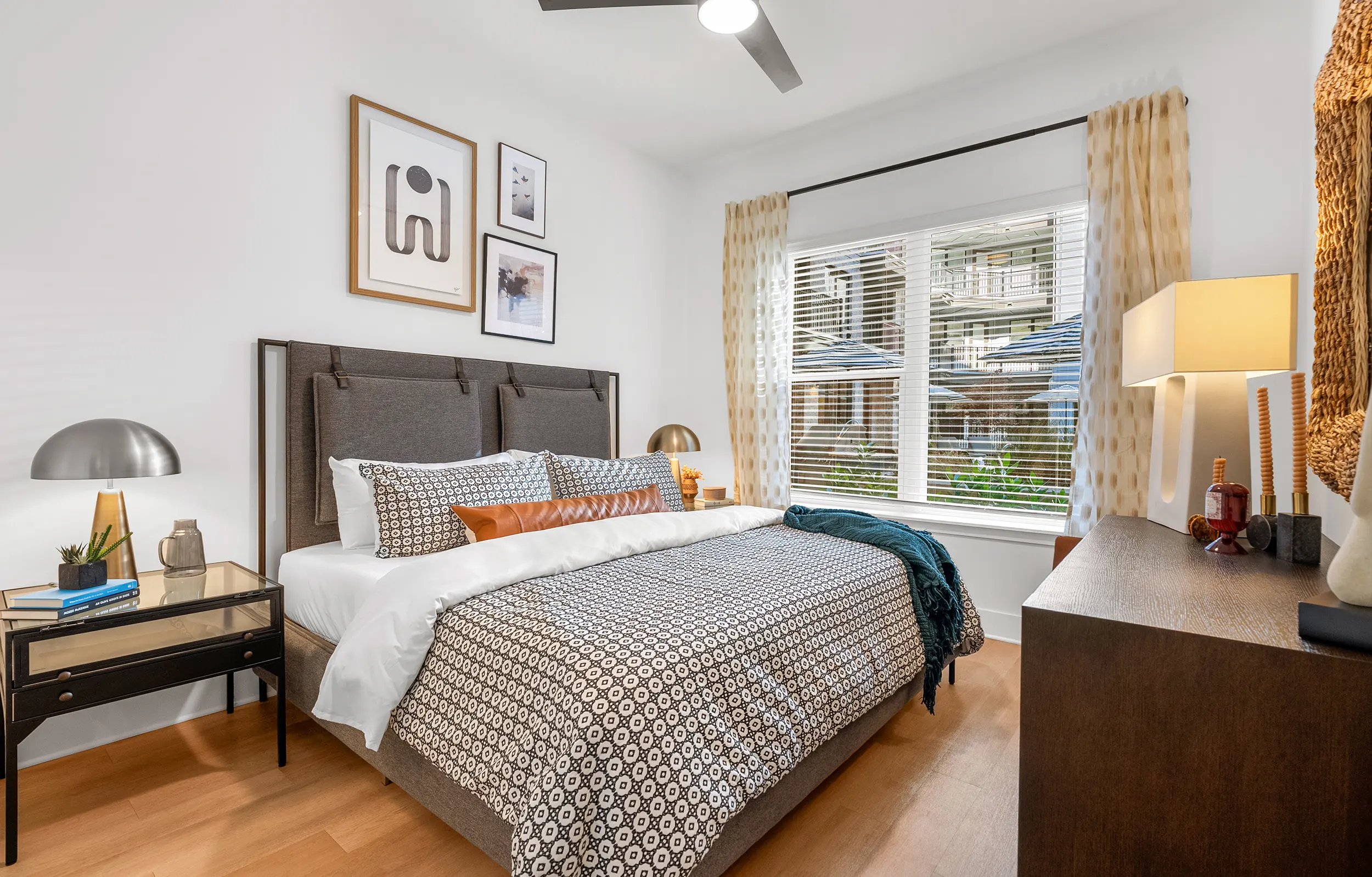 A modern bedroom featuring a queen-sized bed with patterned bedding, a bedside lamp, and a small nightstand. Bright window light enters through sheer curtains, illuminating the room with decorative wall art above the headboard and a wooden dresser nearby.