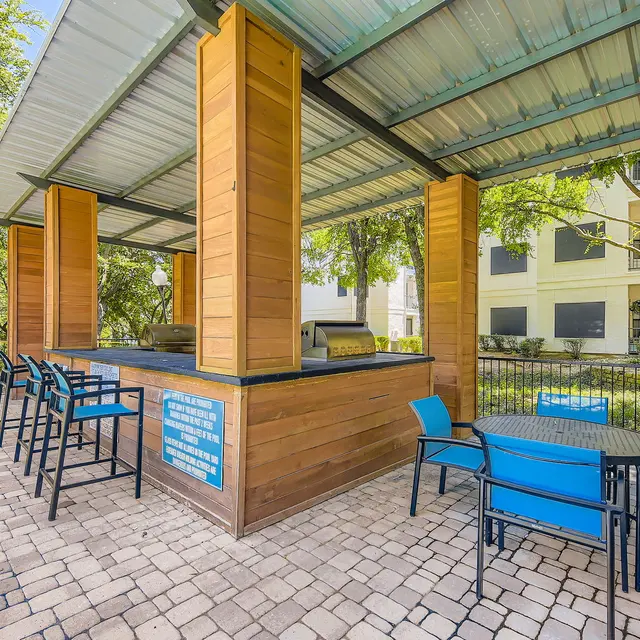 Outdoor kitchen area with a bar and seating under a roof, surrounded by trees and buildings.