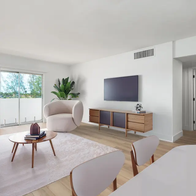 A bright and modern living room featuring a light-colored sofa, a round coffee table, and a large television mounted on the wall. There are indoor plants for decoration and a white rug on the wooden floor, with a balcony visible in the background.
