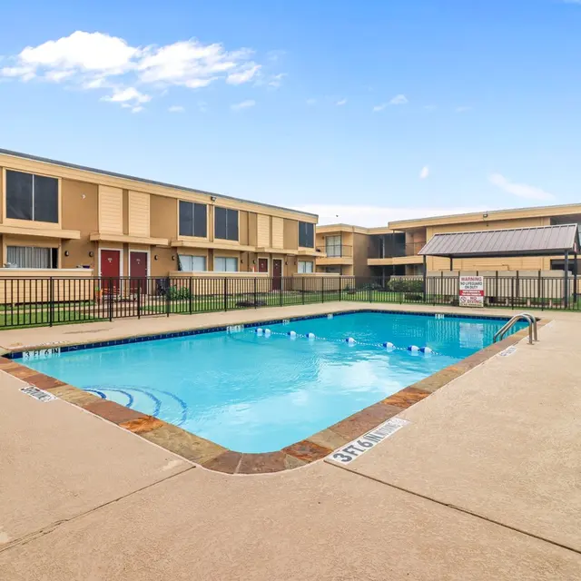 A swimming pool surrounded by a concrete deck and a black fence, with apartment buildings in the background and a clear blue sky above.