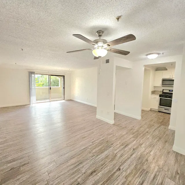 A spacious living room featuring a ceiling fan, a sliding glass door leading to a small balcony, and a kitchen area visible in the background. The floor is made of light wooden planks, and the walls are painted in a neutral color.