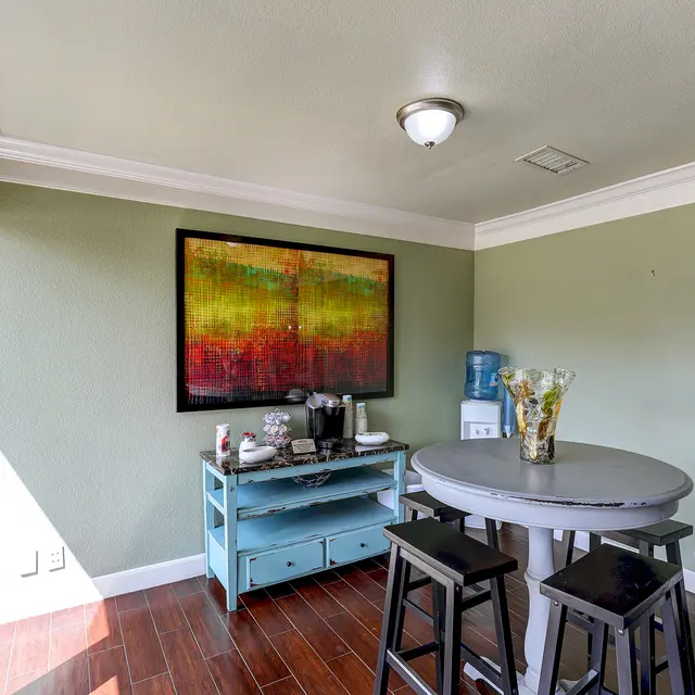 A cozy dining area featuring a small round table with high stools, a water dispenser, and a colorful piece of abstract artwork on the wall.