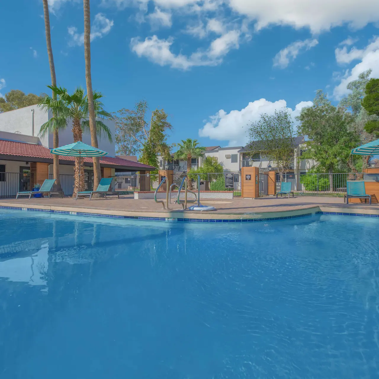 A clear blue pool area surrounded by lounge chairs and umbrellas, framed by palm trees and a blue sky with fluffy clouds.