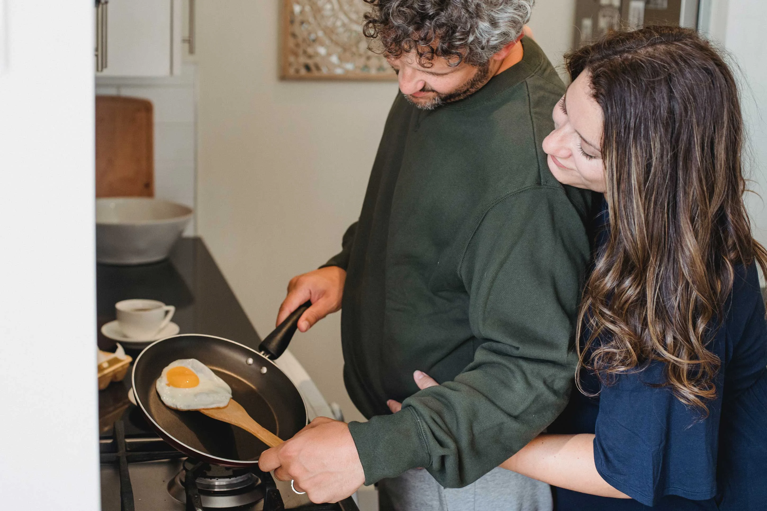 A couple in a cozy kitchen, with one person leaning in from behind while the other flips an egg in a frying pan.