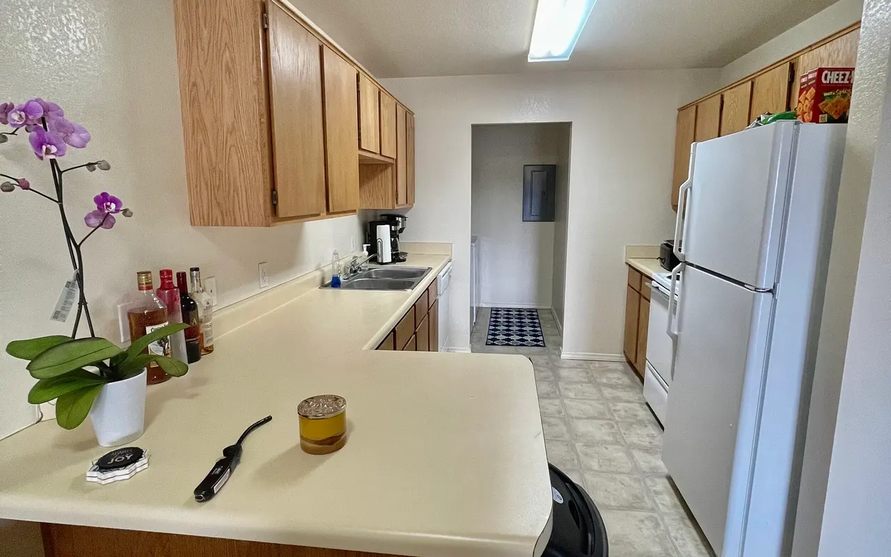 A view of a modern kitchen featuring wooden cabinets, a countertop with a drink, and a trash can. There is kitchen equipment and a refrigerator visible. The floor has a light-colored tile design.
