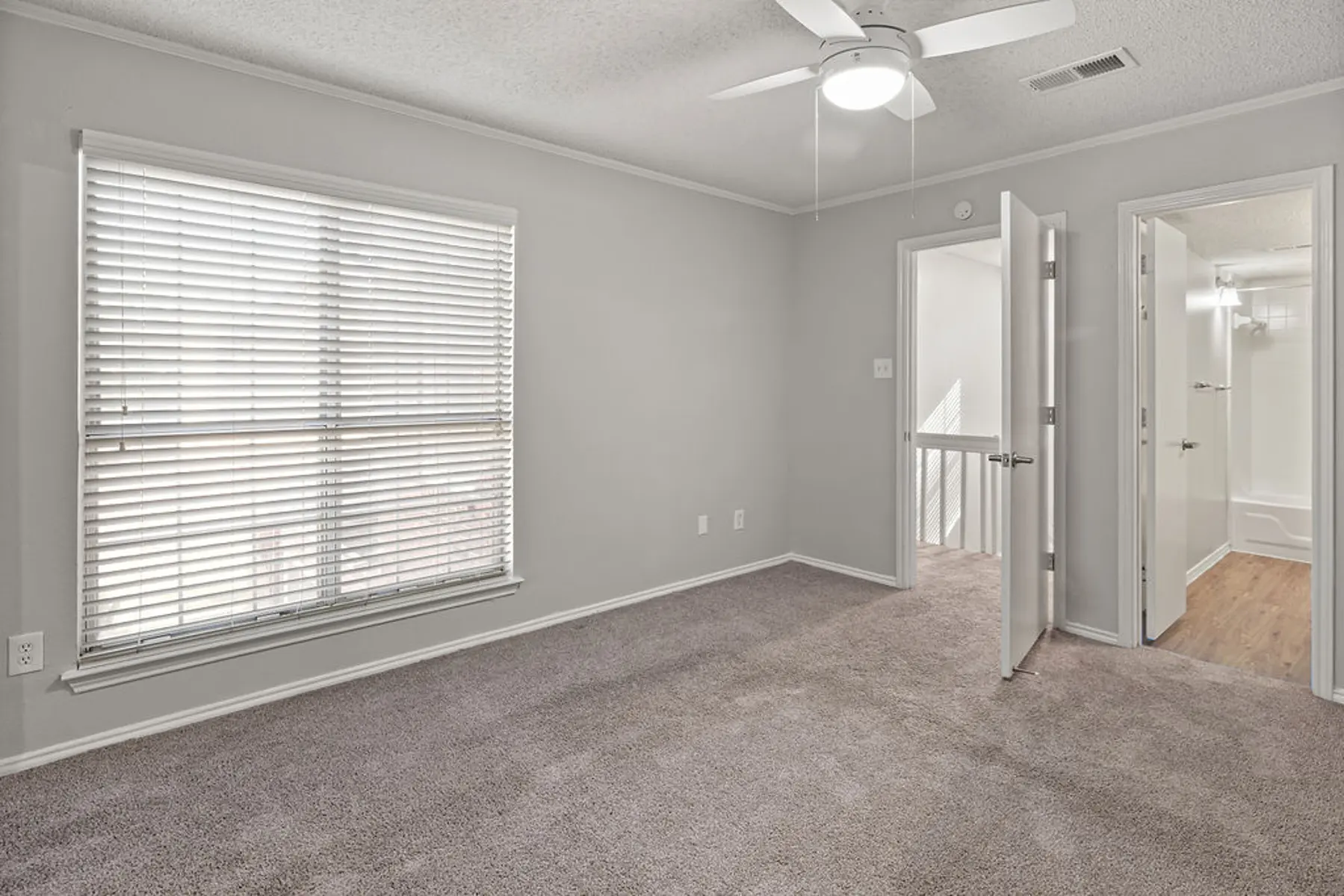 Empty Room with Window and Ceiling Fan A vacant room with light gray walls and carpet, featuring a window with white blinds and a ceiling fan. A partially open door leads to another space, indicating the layout of the area.