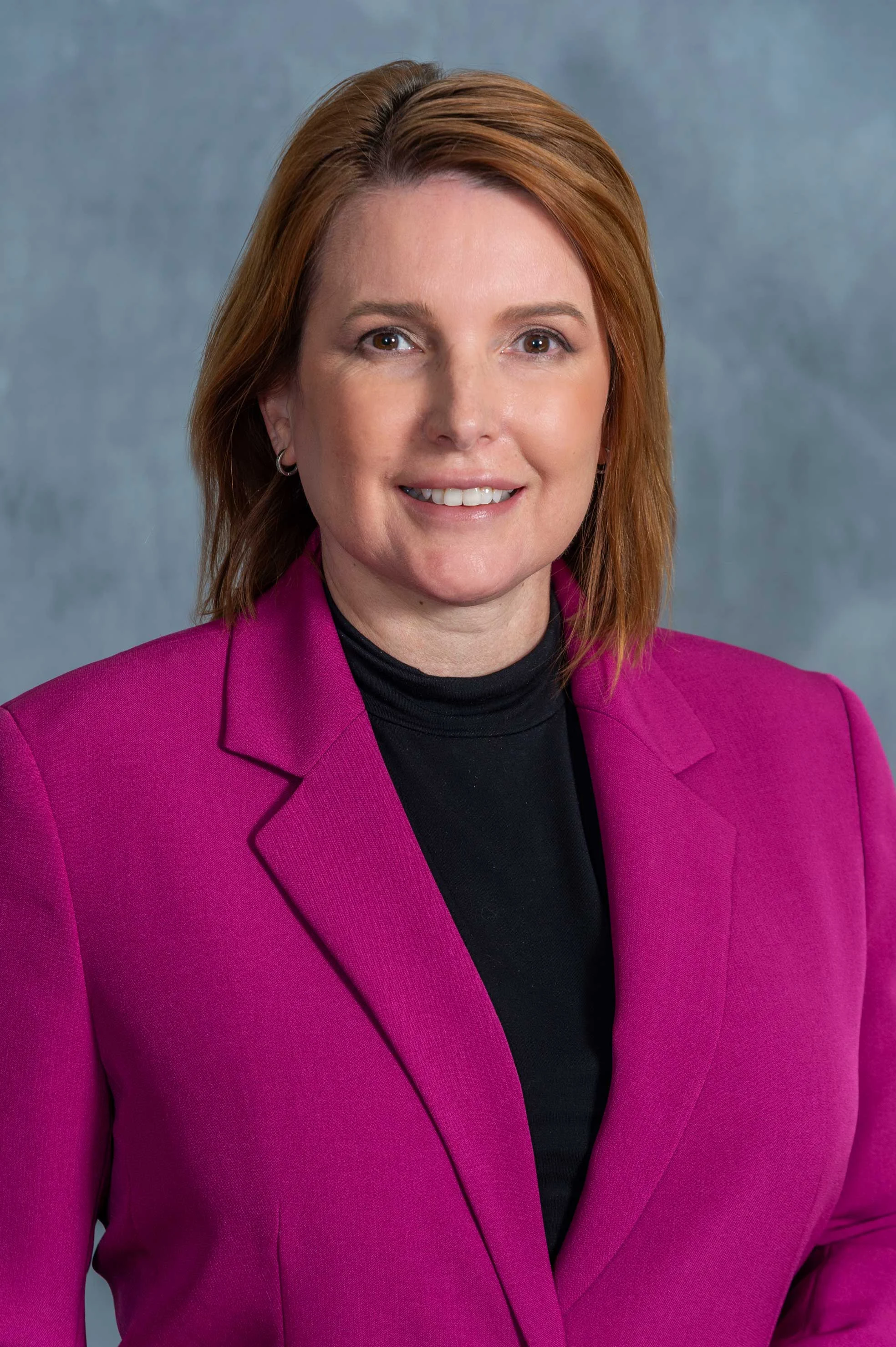A professional woman with shoulder-length hair wearing a bright pink blazer over a black turtleneck. She has a friendly smile and is posed against a simple gray background.