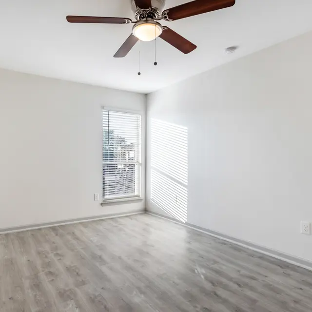 An empty bedroom with light wood floors and a ceiling fan. A window with blinds allows natural light to enter, casting shadows on the floor.