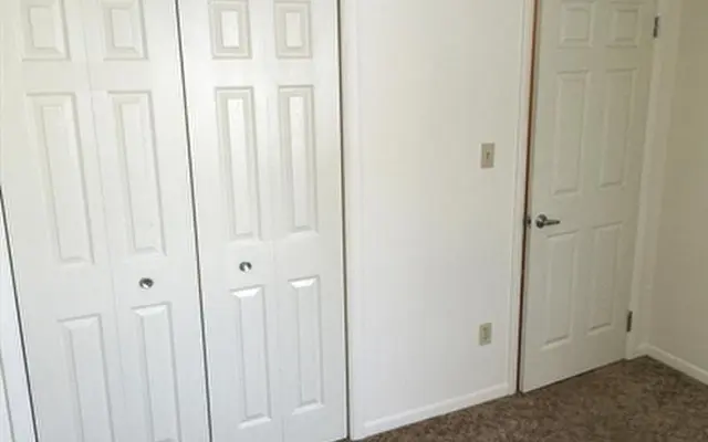 An empty room featuring two white bi-fold closet doors on the left and a single white door on the right, with beige carpet flooring and light-colored walls.