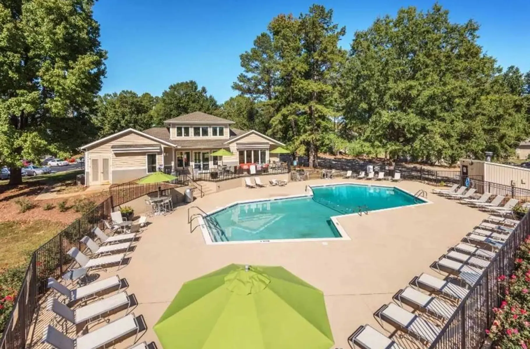 A spacious community pool area featuring a rectangular swimming pool surrounded by lounge chairs and umbrellas, with a clubhouse in the background. Lush green trees provide shade around the area.