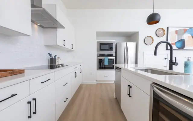 A modern kitchen featuring white cabinetry, stainless steel appliances, and minimalist decor. The island is equipped with a sink and sleek faucet, with light wood flooring complementing the bright ambiance.