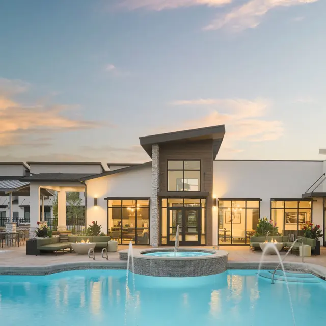 A modern pool area featuring clear turquoise water with fountains, surrounded by lounge chairs and greenery. A stylish building with large windows and a contemporary design stands in the background, under a soft sunset sky.
