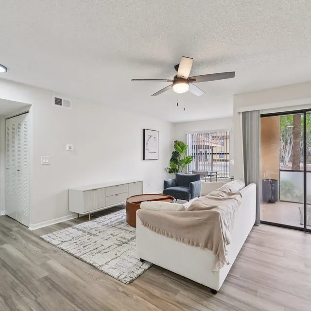 A modern living room with a cozy sofa, ceiling fan, and large sliding glass door leading to a balcony. The decor includes a light rug and a wooden coffee table.