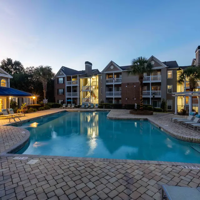 A spacious apartment complex pool area at sunset, featuring a large swimming pool surrounded by palm trees, lounge chairs, and well-lit buildings in the background.