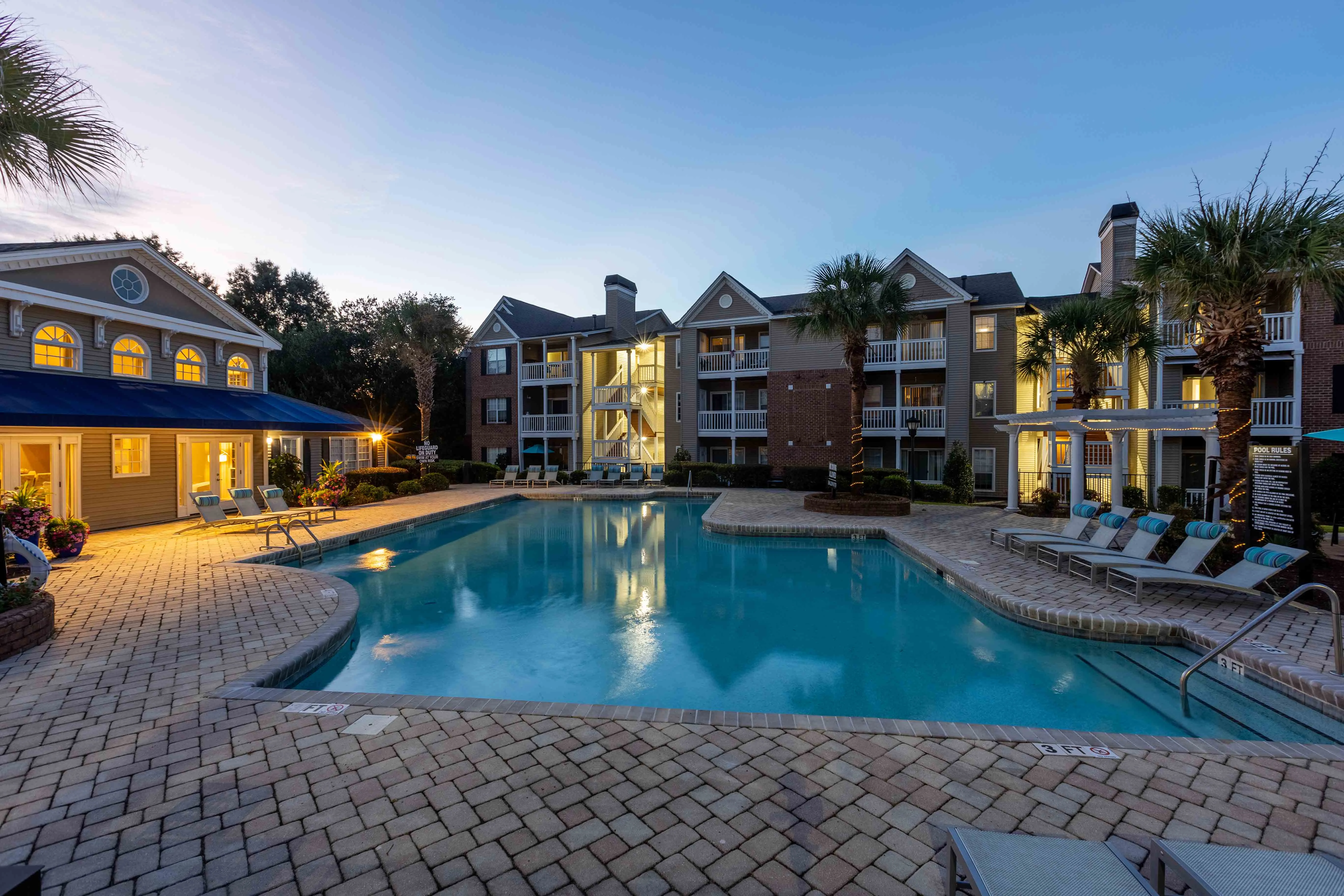 Modern Apartment Pool Area at Sunset A spacious apartment complex pool area at sunset, featuring a large swimming pool surrounded by palm trees, lounge chairs, and well-lit buildings in the background.