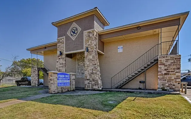 Exterior view of the Spanish Cove Apartments, a two-story building with a stone facade and staircase leading to the upper level.