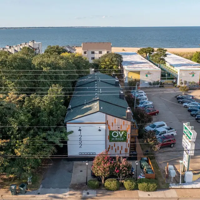 An aerial view of a building near the shoreline, surrounded by trees and a parking area with several cars. The ocean can be seen in the background under a clear sky.