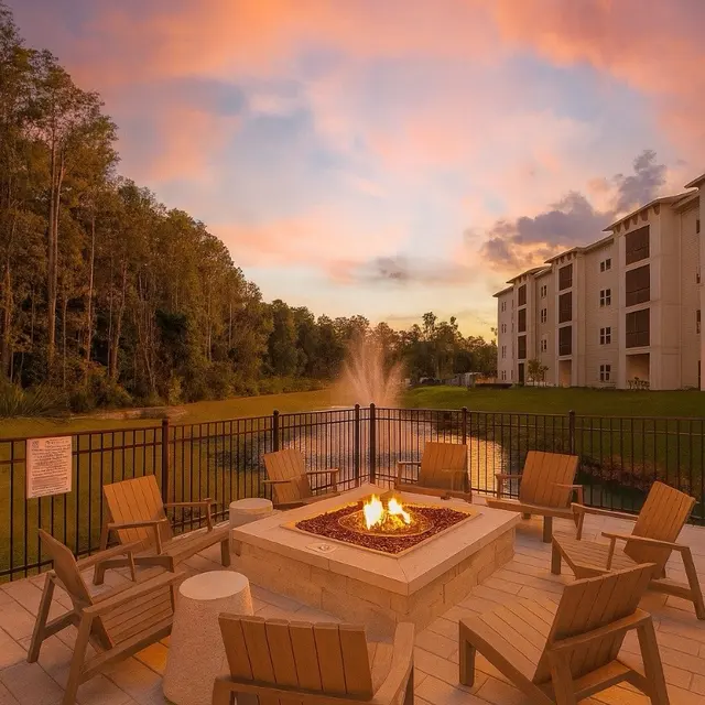 An outdoor fire pit area with wooden chairs, surrounded by a fence and overlooking a pond, under a colorful sunset sky.